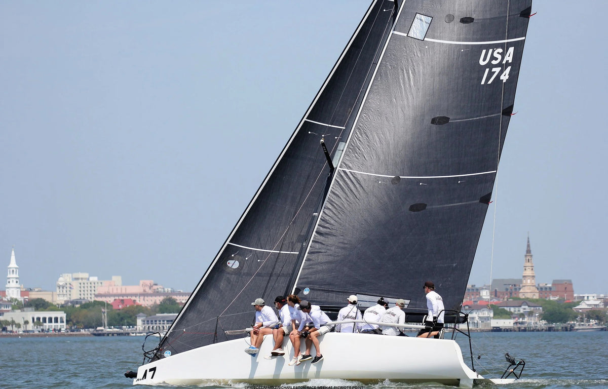 A white racing sailboat with a large black mainsail glides across the water under a clear sky. Several crew members are seated on the deck, and a city skyline is visible in the background.